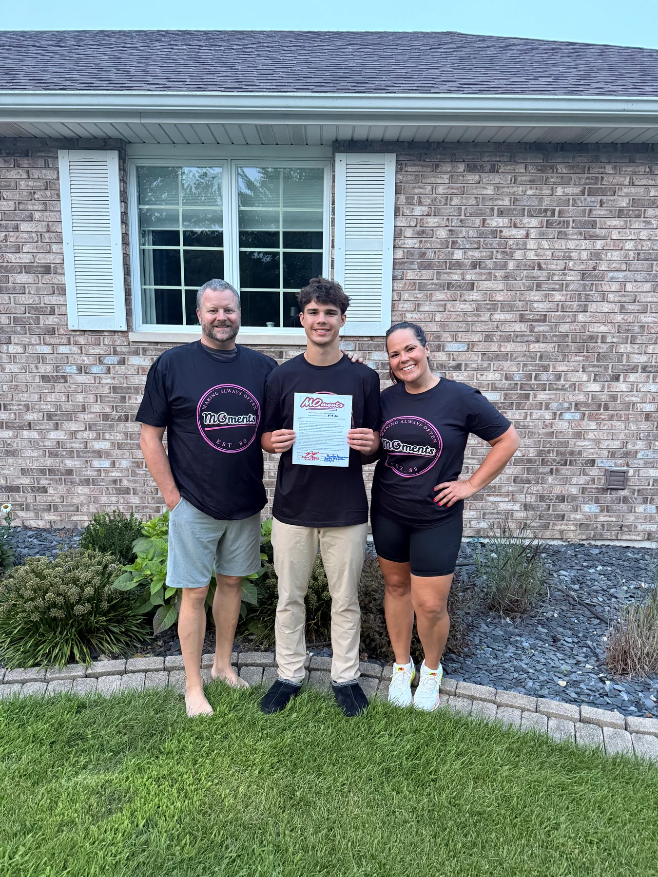 An athlete and two supporters wear matching MOments shirts outside a brick house